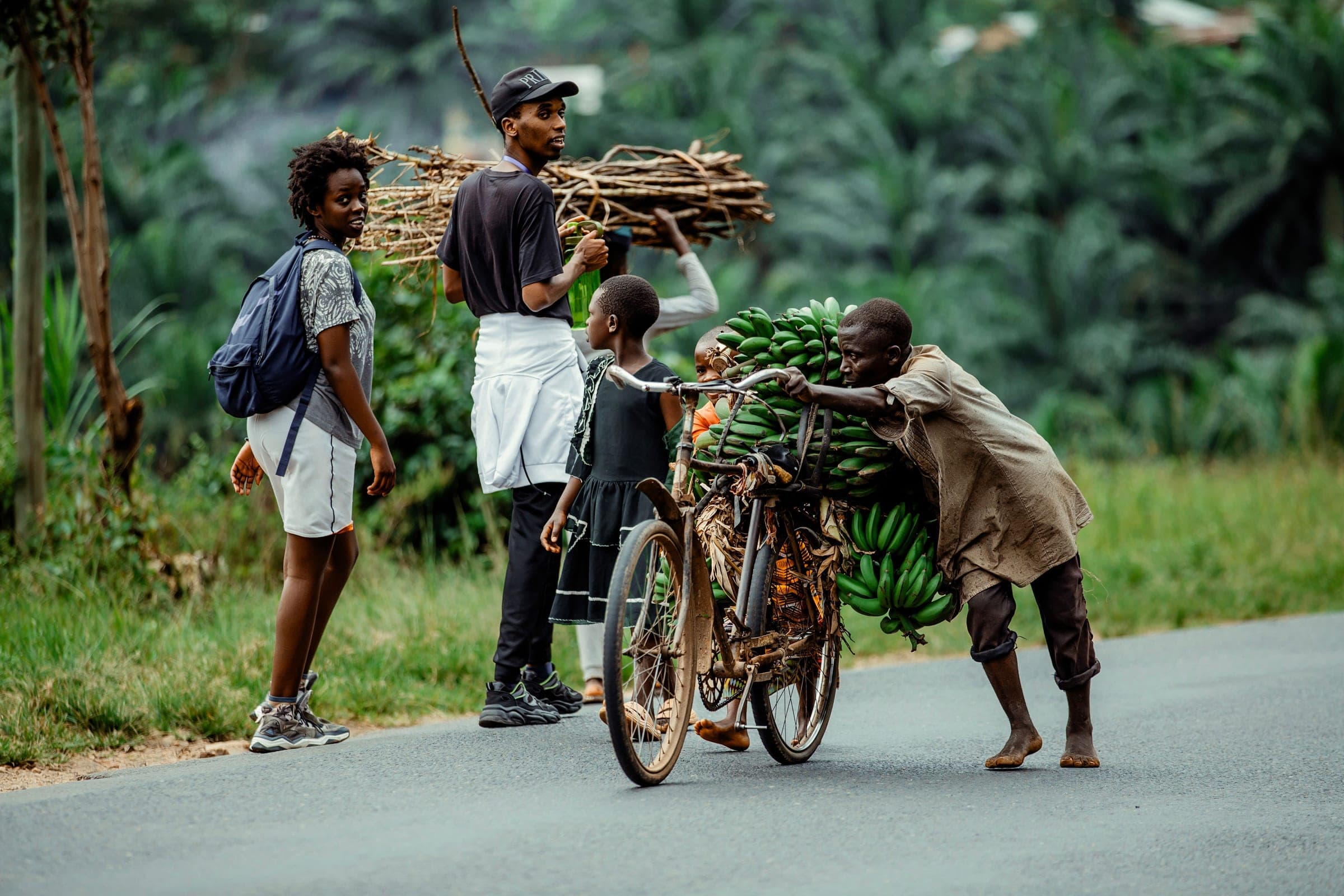Villagers pushing a bicycle loaded with bananas and firewood along a road